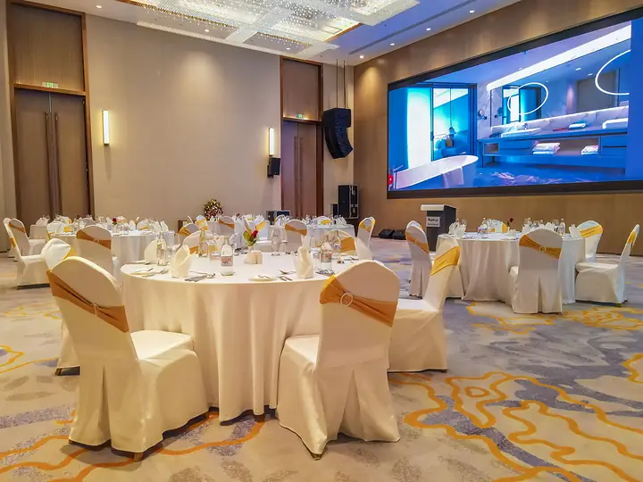 Hall set for a banquet: round tables in white linen with gold chair sashes beside a large LED wall